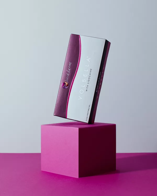 En rektangulär låda med Juvederm Volbella med Lidocaine står upprätt på en rosa kub. Lådan är vit med lila accenter och text, mot en slät bakgrund med skugga.