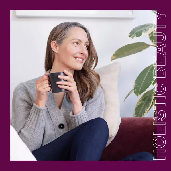 En kvinna med långt brunt hår sitter i en soffa med en mugg i handen, klädd i grå tröja och mörka byxor. Hon ler och tittar åt sidan. En växt och en kudde finns i bakgrunden. Orden "HOLISTIC BEAUTY" står till höger.