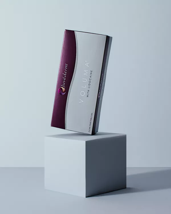 En låda med Juvederm Voluma med Lidocaine står upprätt på en vit kub mot en ljusgrå bakgrund. Lådan har rödbruna och vita delar med synligt produktvarumärke.