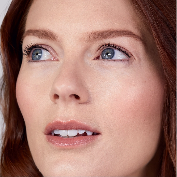 Närbild av en kvinna med ljus hy, blå ögon och långt rödbrunt hår. Hon tittar något uppåt och åt sidan. Hennes makeup är naturlig med markerade ögonbryn, mascara och nudefärgat läppstift.